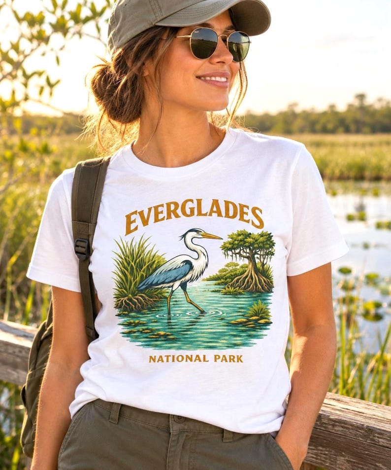 May include: A white short-sleeved t-shirt featuring a graphic print of a blue and white heron standing in water with green marsh grass and a cypress tree. The text EVERGLADES is printed in gold above the bird and NATIONAL PARK below it.
