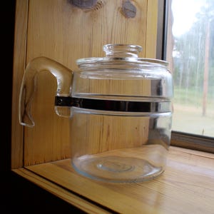 Könnte beinhalten: Eine Kaffeekanne aus klarem Glas mit Griff und Deckel, mit einem schwarzen Metallband in der Mitte. Die Kanne steht auf einer Holzoberfläche in der Nähe eines Fensters.