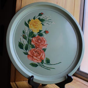Puede incluir: Una bandeja redonda de color verde claro con un diseño floral. La bandeja presenta un grupo de rosas amarillas y coral con hojas verdes. La bandeja se exhibe sobre un soporte negro y está cerca de una ventana.