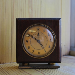 May include: A vintage, square clock in a dark wood case. The clock face is a pale yellow with black numerals and hands. It rests on a small, rectangular base with brass-coloured feet. The clock is likely from the mid-20th century. The clock measures approximately 10 cm in height.