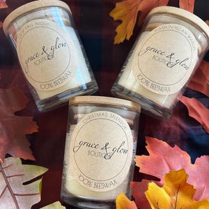 Puede incluir: Tres velas en tarros de vidrio con tapas de madera, cada una etiquetada con un aroma diferente: "Crisp Fall Night", "Pumpkin Pecan Waffles" y "Christmas Splendor". Las etiquetas dicen "grace & glow" y "100% beeswax". Las velas están sobre un fondo a cuadros con hojas de otoño.