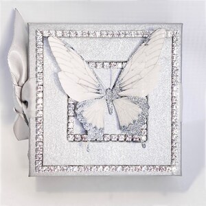 May include: A silver glitter box with a white butterfly design. The box is decorated with a silver rhinestone border.