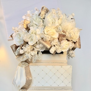 May include: A white wedding card box with a gold trim and a white floral arrangement on top. The box is decorated with rhinestones and has a satin ribbon.