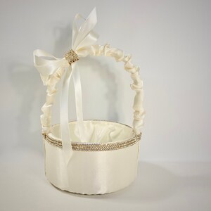 May include: A white satin flower girl basket with a ruffled handle and a large white satin bow with a rhinestone embellishment. The basket is decorated with a band of rhinestones around the top.