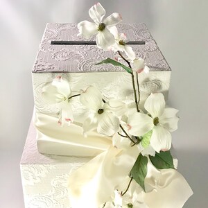 May include: White wedding card box with a white satin ribbon and white flowers. The box has a decorative embossed pattern.
