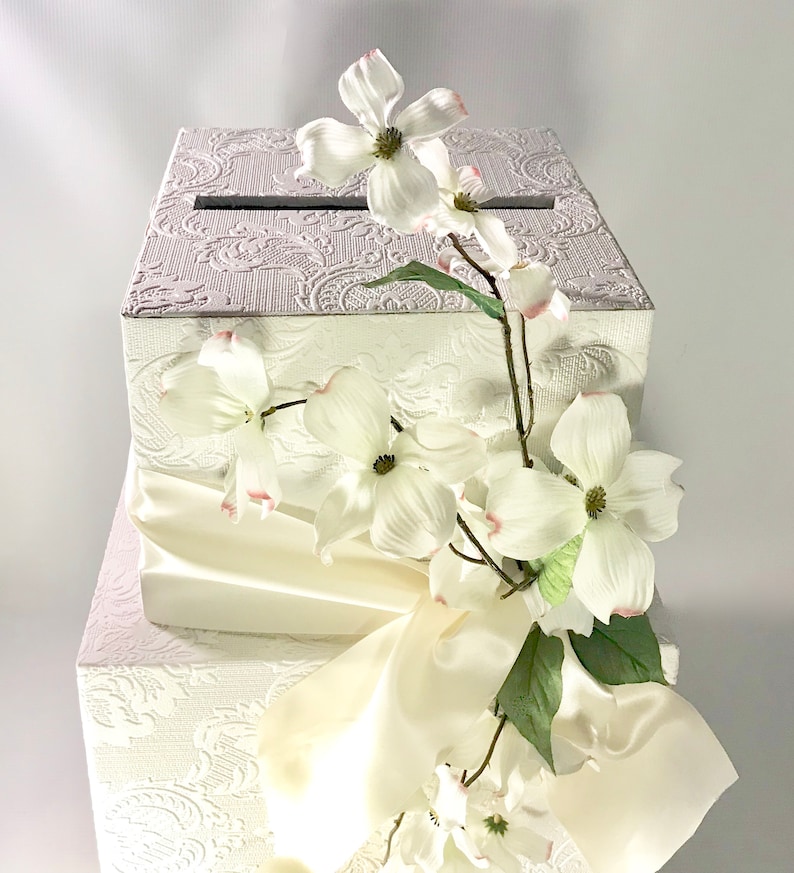May include: White wedding card box with a white satin ribbon and white flowers. The box has a decorative embossed pattern.