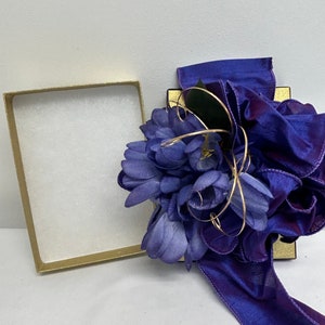 May include: A purple gift box with a purple satin ribbon and a purple flower arrangement. The box is open and the inside is lined with white paper.