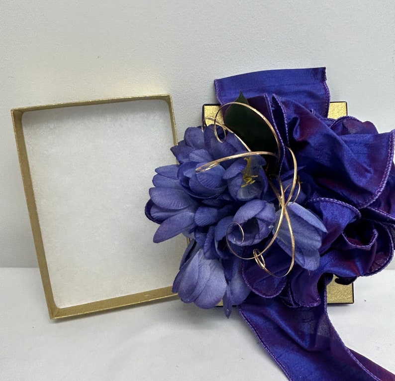 May include: A purple gift box with a purple satin ribbon and a purple flower arrangement. The box is open and the inside is lined with white paper.
