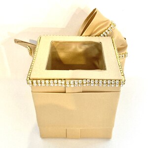 May include: A beige tissue box cover with a clear plastic window and a gold trim decorated with rhinestones.