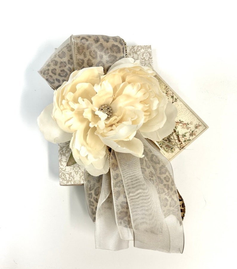 Puede incluir: Una caja de regalo envuelta en papel de estampado de leopardo beige y marr&oacute;n con una gran flor blanca y una cinta beige.