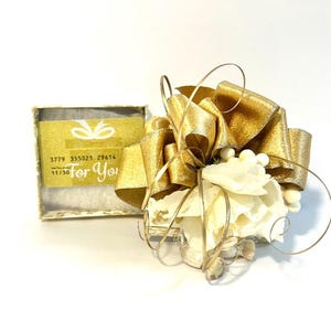 May include: A small white gift box with a gold ribbon and bow. The box is decorated with white flowers and seashells. The box has a gift card inside with the text "For You" and a gift card number.