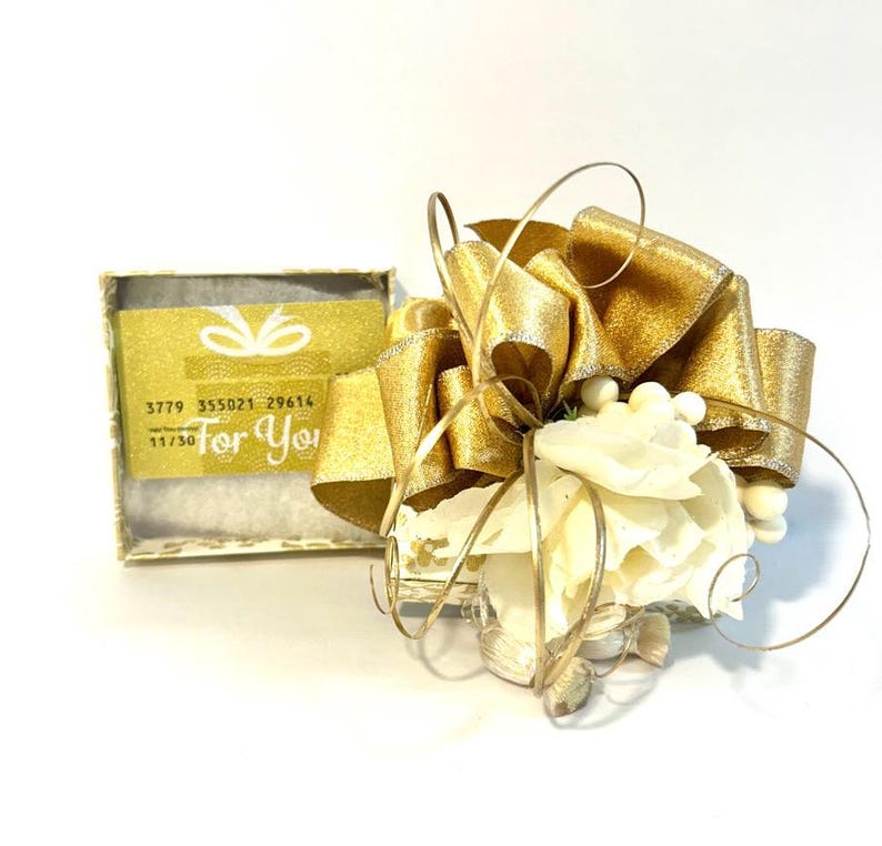 May include: A small white gift box with a gold ribbon and bow. The box is decorated with white flowers and seashells. The box has a gift card inside with the text "For You" and a gift card number.