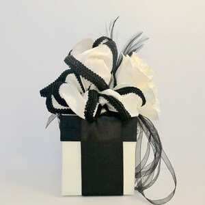 May include: A white gift box with a black ribbon and a white bow with black trim. The bow is decorated with a black feather.