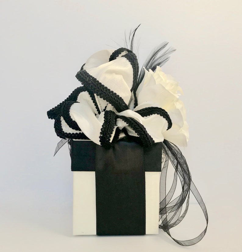 May include: A white gift box with a black ribbon and a white bow with black trim. The bow is decorated with a black feather.
