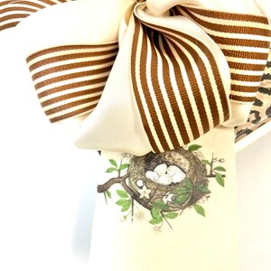 May include: A white and brown striped bow with a small rhinestone embellishment. A white card with a brown bird's nest and white eggs is attached to the bow.