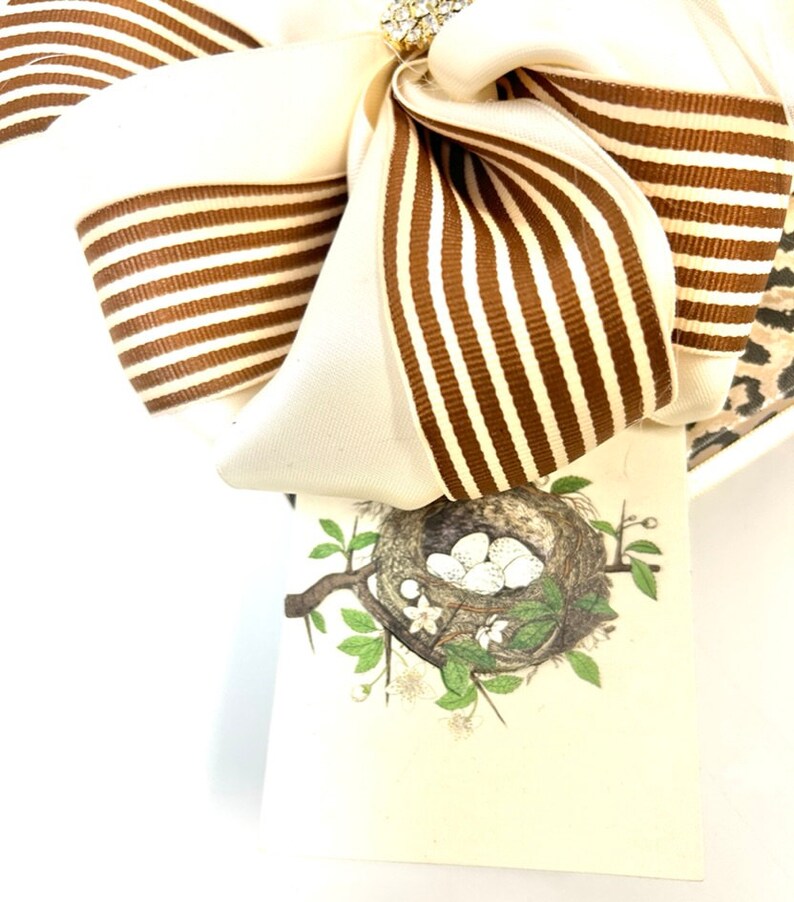 May include: A white and brown striped bow with a small rhinestone embellishment. A white card with a brown bird's nest and white eggs is attached to the bow.