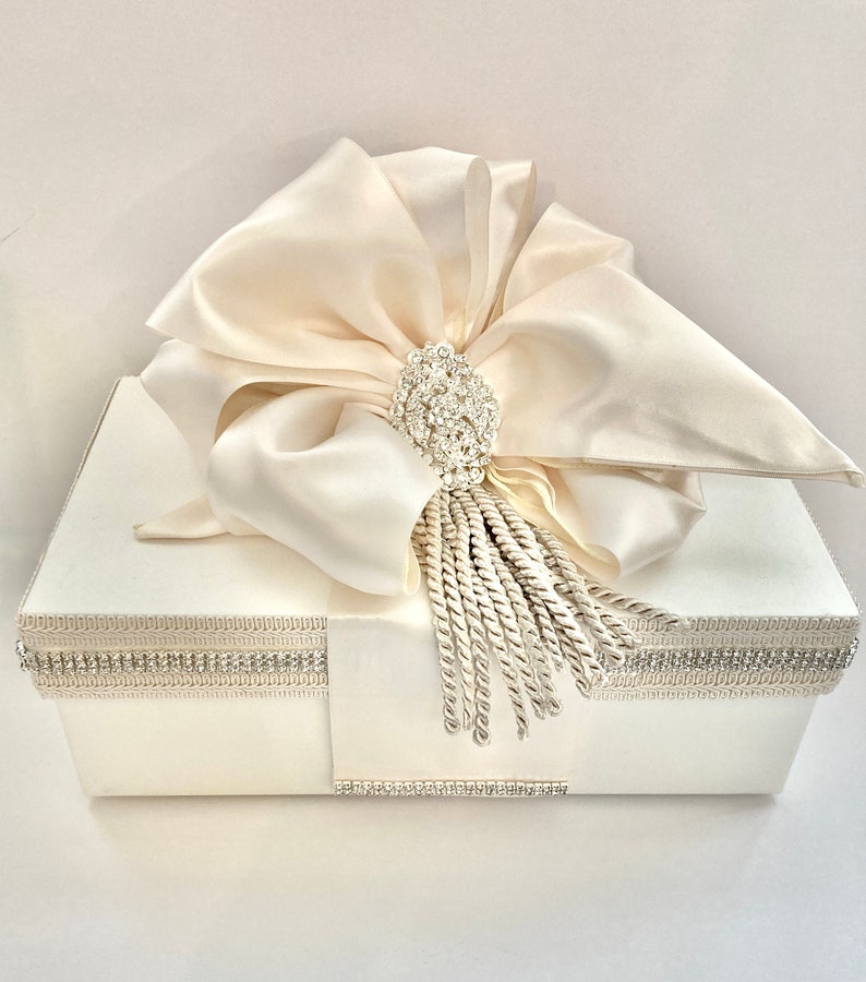 May include: Ivory-colored gift box with a large satin bow and a sparkling rhinestone brooch. The box is trimmed with a rhinestone border and a decorative tassel, creating an elegant and luxurious presentation.