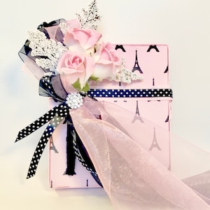 K&ouml;nnte beinhalten: Eine rosa Geschenkbox mit schwarzem Eiffelturm-Muster, verziert mit rosa Rosen, wei&szlig;en Zweigen und schwarz-wei&szlig;em Polka-Dot-Band. Eine glitzernde Brosche und ein durchsichtiges rosa Band sorgen f&uuml;r Eleganz.