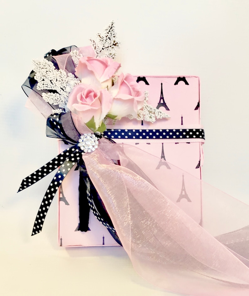 K&ouml;nnte beinhalten: Eine rosa Geschenkbox mit schwarzem Eiffelturm-Muster, verziert mit rosa Rosen, wei&szlig;en Zweigen und schwarz-wei&szlig;em Polka-Dot-Band. Eine glitzernde Brosche und ein durchsichtiges rosa Band sorgen f&uuml;r Eleganz.