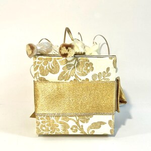May include: A white box with a gold floral pattern and a gold ribbon. The box has a gold clasp and is decorated with white feathers.