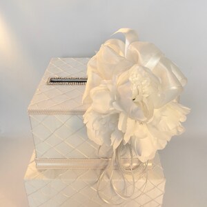 May include: White wedding card box with a white flower and ribbon decoration. The box has a quilted design and a slot for cards.