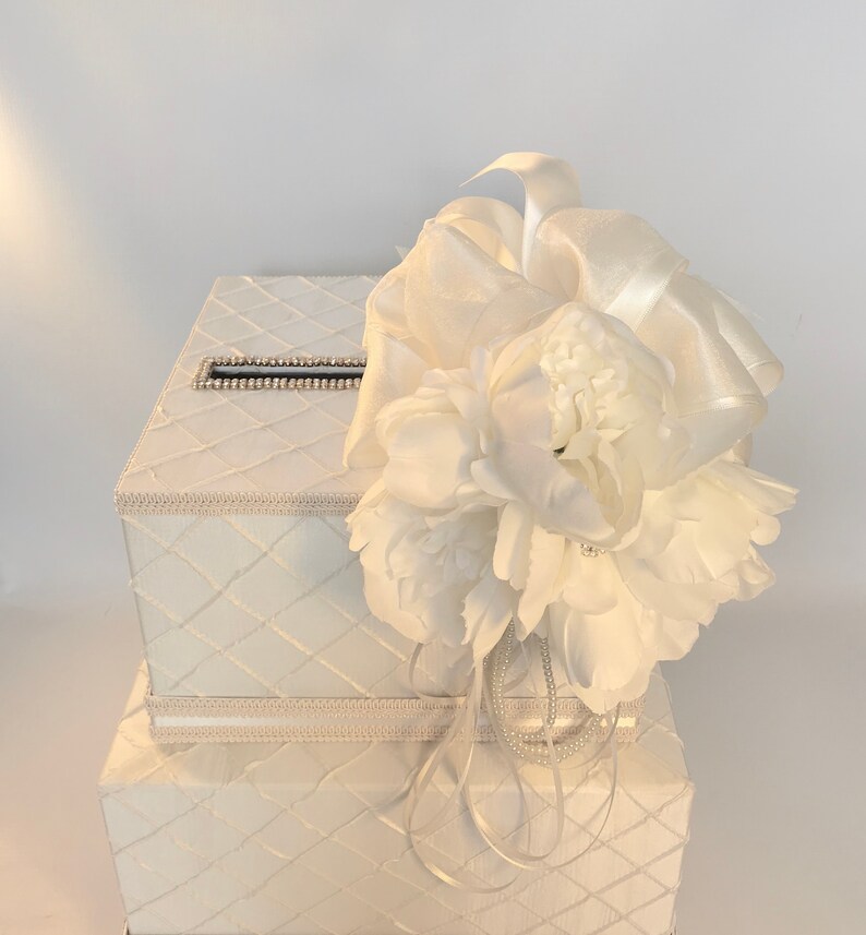 May include: White wedding card box with a white flower and ribbon decoration. The box has a quilted design and a slot for cards.