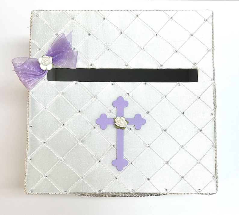 May include: White satin box with a purple cross and a bow with white flowers. The box has a slot for cards or envelopes.