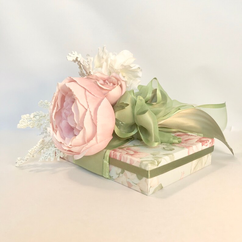 K&ouml;nnte beinhalten: Eine gr&uuml;ne und wei&szlig;e Blumen-Geschenkbox mit einem rosa und wei&szlig;en Blumenstrau&szlig;, der mit einem gr&uuml;nen Band gebunden ist.
