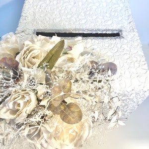 May include: White wedding card box with a lace design and a clear plastic slot for cards. The box is decorated with a bouquet of white roses and greenery.