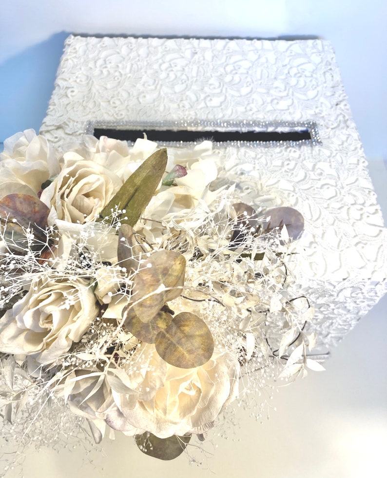 May include: White wedding card box with a lace design and a clear plastic slot for cards. The box is decorated with a bouquet of white roses and greenery.