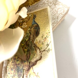Puede incluir: Una postal vintage con un pavo real con la cabeza azul y plumas verdes y marrones, posado en una rama con flores rosas. El fondo es un patr&oacute;n dorado y blanco.