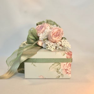 Puede incluir: Una caja de regalo blanca con un dise&ntilde;o floral con rosas rosas y hojas verdes. La caja est&aacute; atada con una cinta de sat&eacute;n verde y rematada con un ramo de rosas rosas, hortensias blancas y aliento de beb&eacute; blanco.