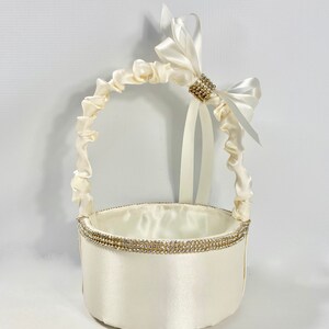 May include: A white satin flower girl basket with a white satin bow and rhinestone embellishment. The basket has a round base and a handle made of gathered satin.