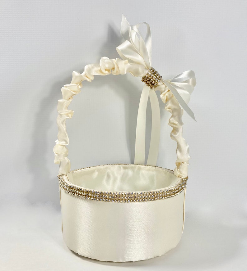 May include: A white satin flower girl basket with a white satin bow and rhinestone embellishment. The basket has a round base and a handle made of gathered satin.