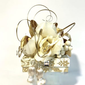 May include: A small white box with a gold and white patterned design. The box is decorated with a white flower arrangement, gold wire, and gold ribbon. The box is topped with two white tassels.