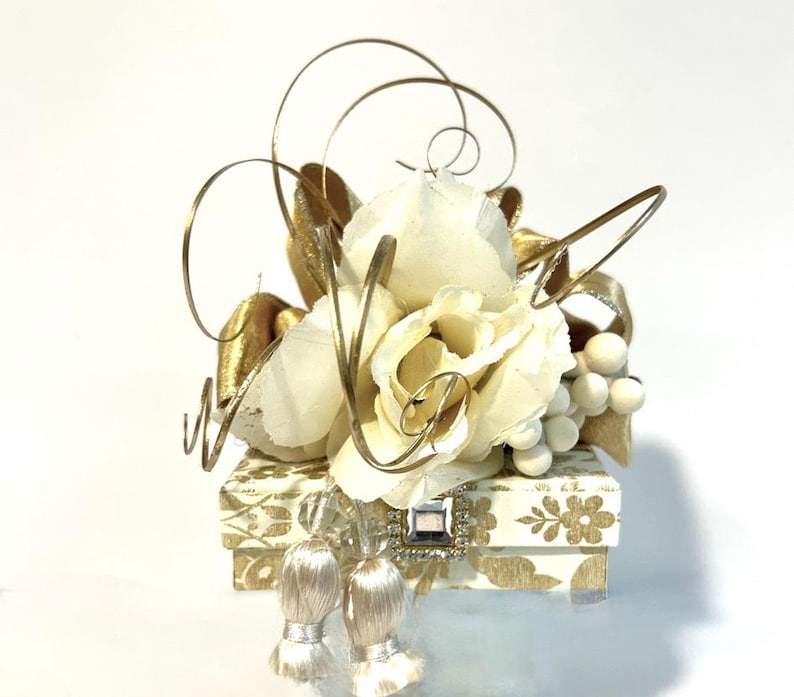 May include: A small white box with a gold and white patterned design. The box is decorated with a white flower arrangement, gold wire, and gold ribbon. The box is topped with two white tassels.