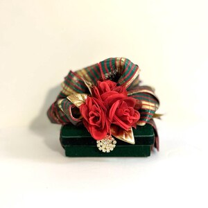 May include: A small green velvet box with a red and gold striped ribbon and three red fabric roses on top. The box has a decorative gold and clear rhinestone embellishment.