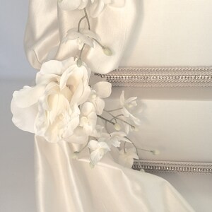 May include: A white cake with a white satin ribbon and white flowers. The cake is decorated with a silver trim.