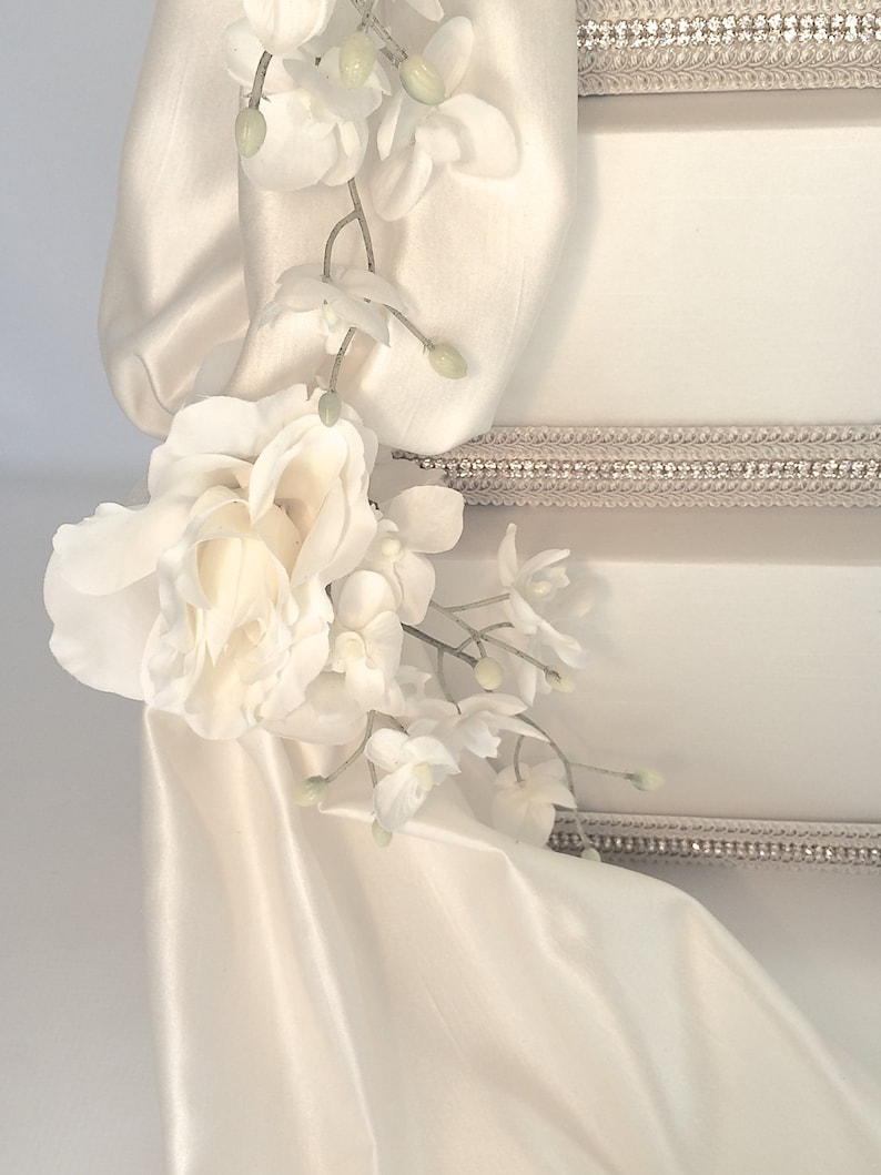 May include: A white cake with a white satin ribbon and white flowers. The cake is decorated with a silver trim.