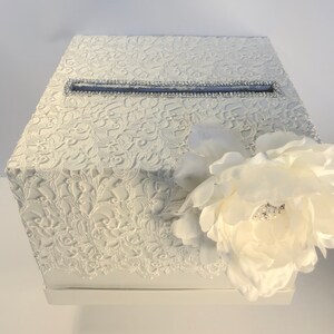 May include: A white, hexagonal card box with a textured lace pattern. A rectangular slot is trimmed with rhinestones. A large white flower with a pearl center adorns the side. Suitable for weddings or special events.