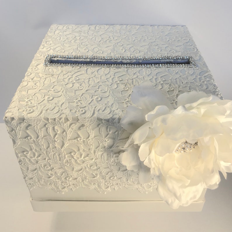 May include: A white, hexagonal card box with a textured lace pattern. A rectangular slot is trimmed with rhinestones. A large white flower with a pearl center adorns the side. Suitable for weddings or special events.