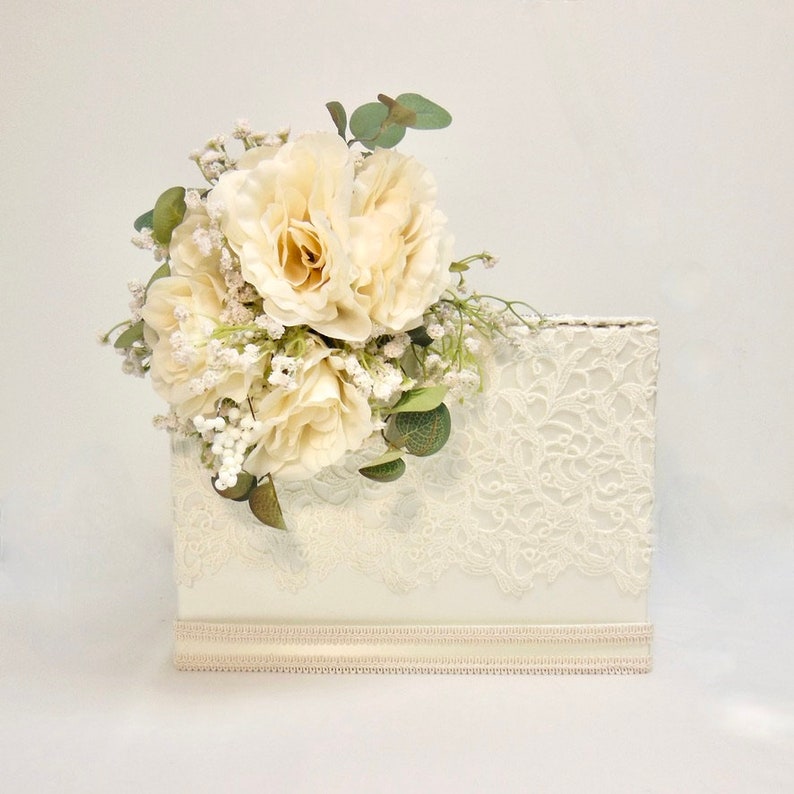 May include: A white lace wedding card box with a bouquet of white roses and greenery on top.