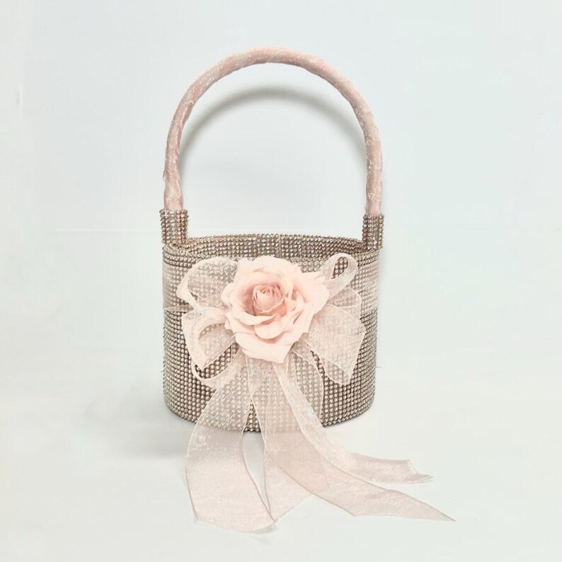 May include: A small, decorative basket with a handle, covered in small, sparkling rhinestones. A large, light pink rose and bow adorn the front. The handle and bow are made of a soft, velvety material. The basket is designed for decorative purposes.