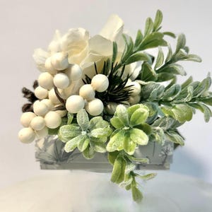 K&ouml;nnte beinhalten: Ein kleiner wei&szlig;er Blumenstrau&szlig; mit wei&szlig;en Beeren, gr&uuml;nen Bl&auml;ttern und einem Hauch von Kiefernnadeln. Die Anordnung befindet sich in einer kleinen grauen Schachtel mit einem wei&szlig;en und grauen Muster.