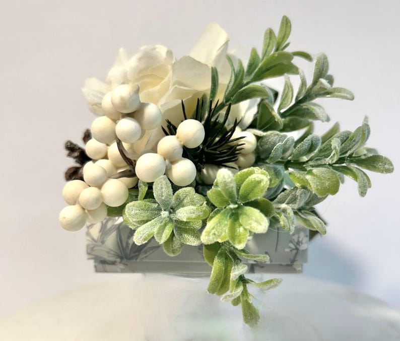 K&ouml;nnte beinhalten: Ein kleiner wei&szlig;er Blumenstrau&szlig; mit wei&szlig;en Beeren, gr&uuml;nen Bl&auml;ttern und einem Hauch von Kiefernnadeln. Die Anordnung befindet sich in einer kleinen grauen Schachtel mit einem wei&szlig;en und grauen Muster.