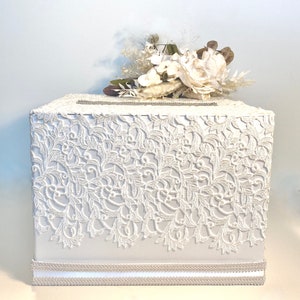 May include: White wedding card box with a lace design and a white floral arrangement on top.