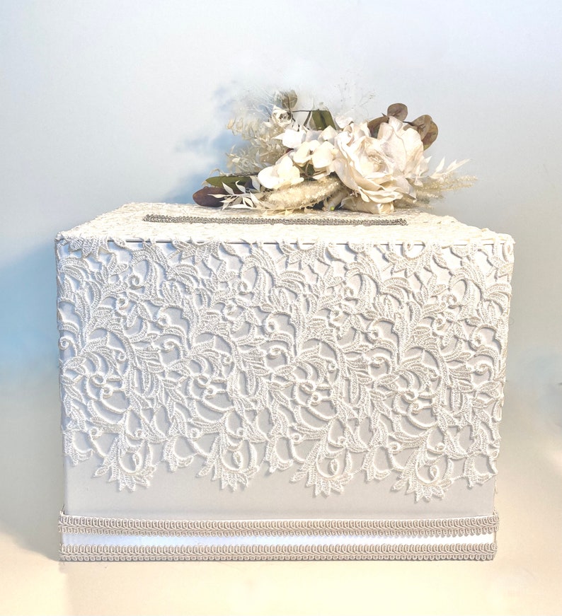 May include: White wedding card box with a lace design and a white floral arrangement on top.