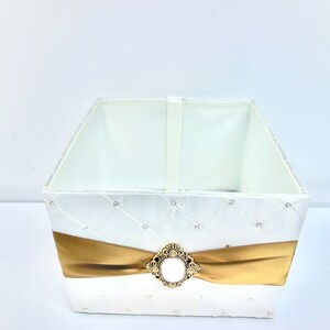 May include: A white square box with a gold ribbon and a gold ornate embellishment. The box is lined with white fabric and has a quilted design.