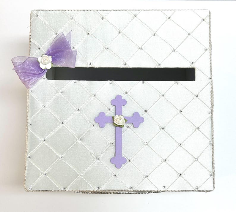 May include: White satin box with a purple cross and a bow with white flowers. The box has a slot for cards or envelopes.