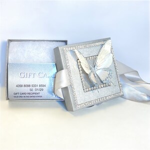 May include: A silver gift card box with a white butterfly design. The box is decorated with rhinestones and has a white satin ribbon. The gift card inside has the text "GIFT CARD" and a gift card number.